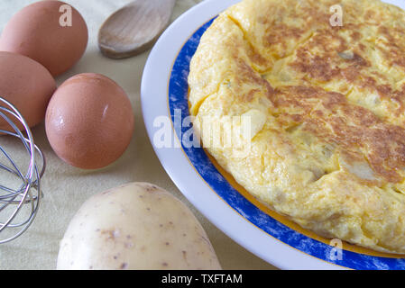 Typical Spanish Tortilla de Patata omelet on a table next to some eggs, potato in a rustic kitchen. Home made plate as a culinaty tourism concept. Stock Photo