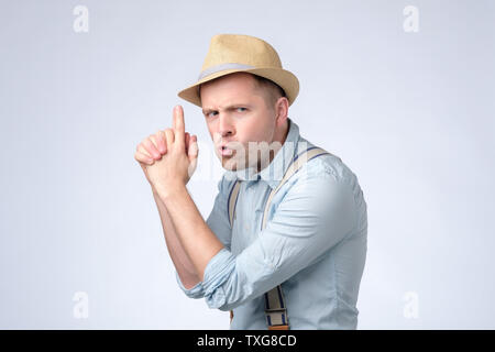 Young handsome man wearing hat and suspenders holding symbolic gun with hand gesture Stock Photo