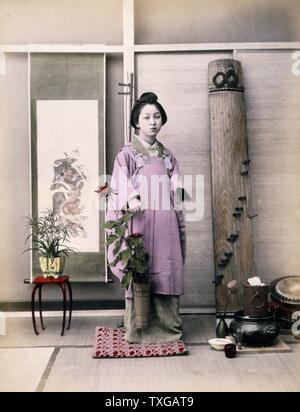 Photograph shows a portrait of a young woman, full-length, standing, facing front, holding a small, long-handled bucket with flowers in it. There is a small table with potted plant in front of a hanging scroll in the background on the left, and a Japanese zither leaning against the wall on the right. Watercolour drawings on the mount depict a Japanese red-crowned crane in flight with Mount Fuji in the background in the upper right and two cranes in the lower left. Stock Photo