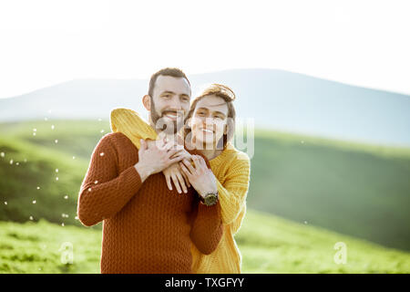 Portrait of a lovely couple dressed in bright sweaters hugging on the green meadow while traveling in the mountains Stock Photo