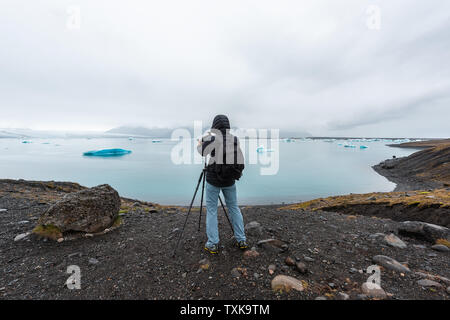 Young man photographer in cold winter coat taking picture photo photographing blue glacier icebergs in lagoon lake in Iceland on tripod Stock Photo