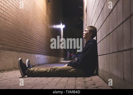 Sad teen sitting in an alleyway all alone at night. Stock Photo
