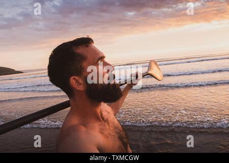 A bearded man walks towards the sea with a Stand Up Paddle board and paddle at sunset on the beach in Devon UK