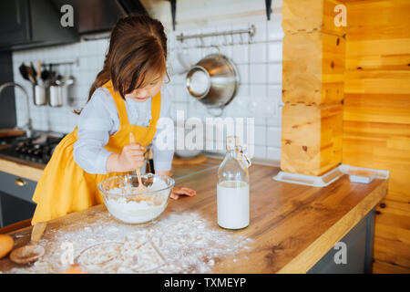 A little cute girl getting ready to bake a cake in the kitchen. Stirring dough in a bowl. Flour and milk on the table. Stock Photo