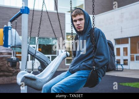 Sad teenager sitting on a swing outside of a school. He is reminiscing about when he was younger.