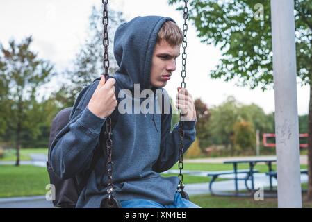 Sad teenager sitting on a swing outside of a school. He is reminiscing about when he was younger. Stock Photo