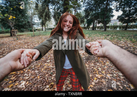 Young woman with long red hair dancing while holding boyfriend's hands in autumn park, personal perspective