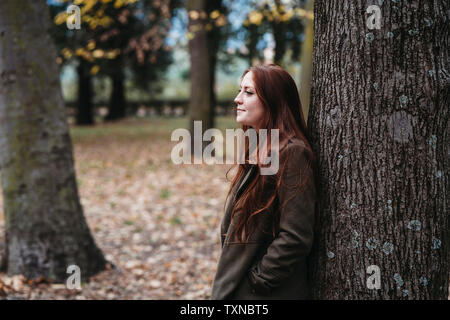 Young woman with long red hair leaning against tree trunk in autumn park
