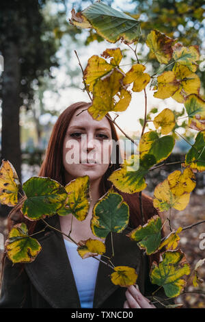 Young woman with long red hair behind twig of autumn leaves in park, portrait Stock Photo