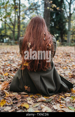 Young woman sitting on leaves in park with autumn leaves tangled in long red hair, rear view