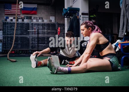 Young woman and man training together in gym, sitting on floor doing warm ups and looking at laptop Stock Photo