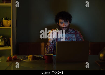 Bearded young man having breakfast, using laptop at home Stock Photo