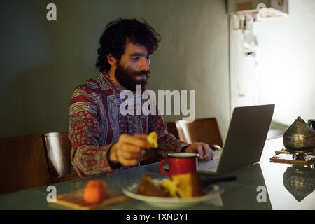 Bearded young man having breakfast, using laptop at home Stock Photo