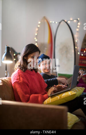 Young woman typing on laptop on living room sofa with young man playing guitar Stock Photo
