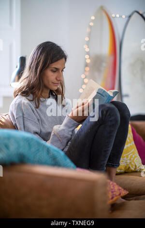 Young woman relaxing on living room sofa reading a book Stock Photo