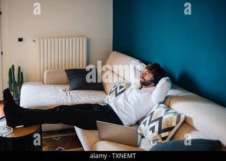 Mid adult man reclining on sofa listening to headphones Stock Photo