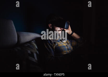 Bearded mid adult man sitting on sofa in darkness looking down through virtual reality headset Stock Photo