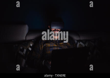 Bearded mid adult man sitting on sofa in darkness looking through virtual reality headset Stock Photo