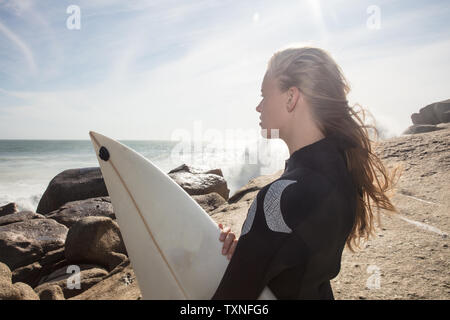 Young female surfer with flyaway hair looking out from beach rock, Cape Town, Western Cape, South Africa Stock Photo