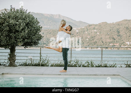Romantic mid adult man lifting up girlfriend on poolside, portrait, Stresa, Piemonte, Italy Stock Photo