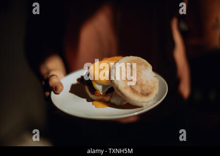 Barista holding plate with fried egg muffin in cafe, close up low key Stock Photo