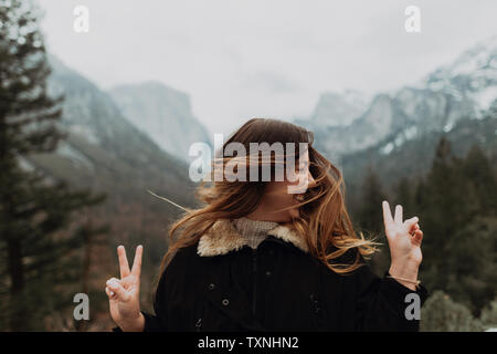 Young woman shaking long brown hair and making peace sign in mountain landscape, Yosemite Village, California, USA Stock Photo