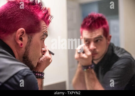 Diverse caucasian male man with spiked pink hair applying eyeliner makeup in mirror's reflection. Wearing black clothes with punk rocker alternative l Stock Photo