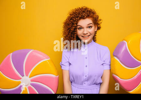 curly red-haired positive woman wearing blue dress with sweet big lollipop on yellow background Stock Photo