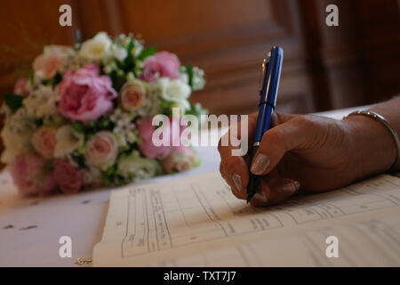 close-up of bride's hand holding pen signing wedding register with floral bouquet in background Stock Photo