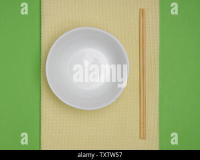 White bowl with asian chopsticks and cupronickel fork and spoon on a yellow towel on green background with copyspace Stock Photo