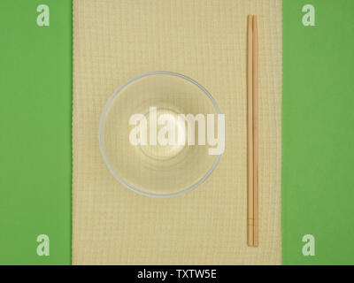 Glass bowl with asian chopsticks and cupronickel fork and spoon on a yellow towel on green background with copyspace Stock Photo