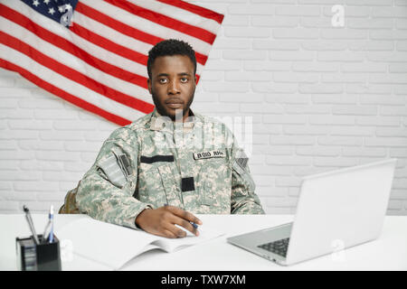 Worker of American army or soldier sitting on table and watching documents, seriously looking at camera. Ranker in uniform making contract to protecting and serving homeland. American flag on wall. Stock Photo