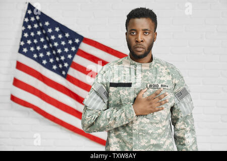 Front view of male American soldier holding hand on heart and seriously looking at camera. Ranker in uniform giving thunderbolt to protecting and serving homeland. American flag on wall. Stock Photo