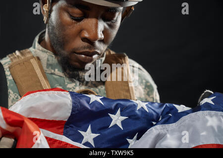 Close up of American soldier in helmet and uniform sadly looking and crying under national flag. Ranker from army holding flag of homeland. Concept of loss and sadness emotions. Stock Photo