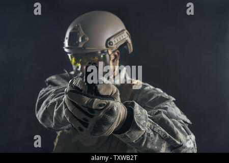 Front view of seriously American soldier in uniform holding modern gun in hands and shooting at camera. Brave ranker wearing helmet keeping position, posing in darkness. Black background. Stock Photo