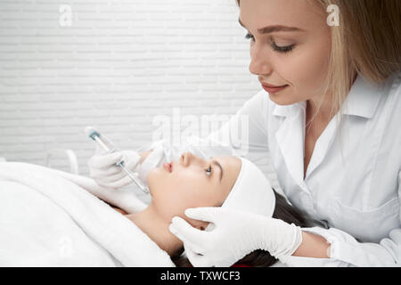 Beautiful woman receiving procedure of correction and beauty in cosmetology cabinet. Client in white bathrobe lying and looking up. Cosmetologist in medical gloves behind making injection on face. Stock Photo