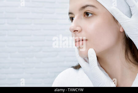 Young, beautiful woman in white bathrobe receiving procedure of inspecting face in cosmetology center. Hands of doctor in white medical gloves touching chin. Concept of skin care. Stock Photo