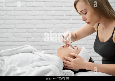 Master of beauty salon holding device and performs procedure with eyelash extension. Young, pretty woman lying on massage table in bathrobe with closed eyes. Concept of beauty. Stock Photo