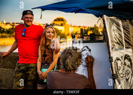 A young couple poses smiling for a portrait that a street artist draws on the bank of the Vltava in Prague, Czechia. In the evening light, a building with a golden roof, possibly the National Theatre, stands in the background. Stock Photo