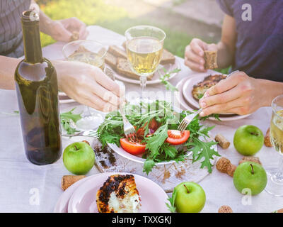 People eat in the garden at the table. Dinner concept with wine in the fresh air. Grilled sea fish and salads with vegetables and herbs. Mediterranean Stock Photo