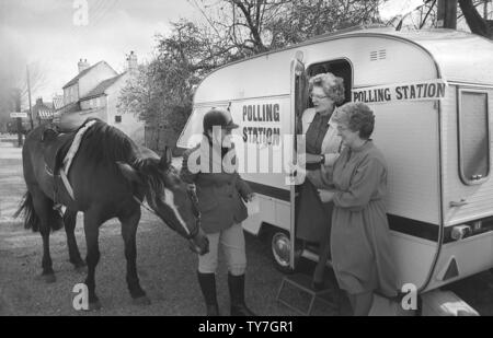 1980s, a lady rider with her horse going to vote at a polling station situated in a small caravan parked in a rural village, Yorkshire, England, UK, being met at the door by two lady presiding officers. Stock Photo