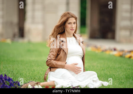 Portrait of happy young pregnant model sitting with crossed legs on grass lawn and looking at her belly with gentle smile. Future mom expecting baby c Stock Photo