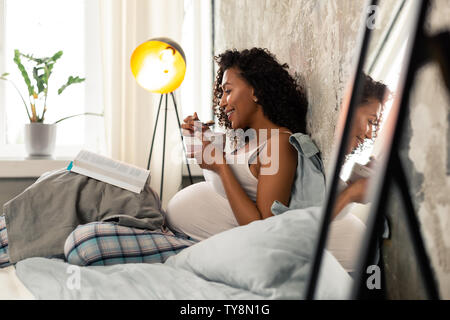 Pregnant woman with a book eating in her bed. Stock Photo