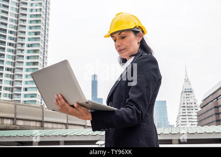 Woman engineer with yellow helmet holding laptop on city background Stock Photo