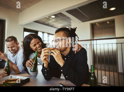 Laughing young African American businesswoman sitting with her arm around a smiling coworker while having pizza and beers together after work Stock Photo