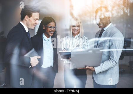 Smiling group of diverse businesspeople standing together in a bright modern office discussing work over a laptop Stock Photo