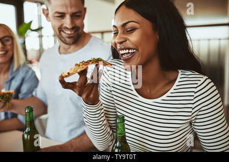 Laughing young African American businesswoman eating pizza and drinking and beer while sitting with colleagues in an office after work Stock Photo