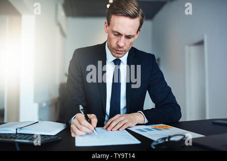 Focused businessman reading paperwork and signing documents while sitting at his desk in a bright modern office Stock Photo
