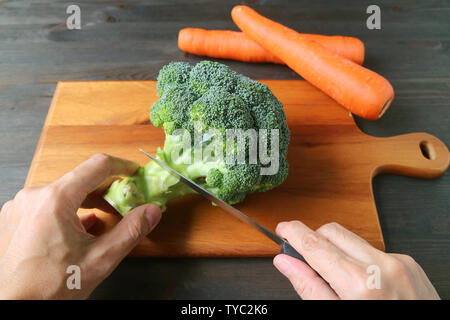 Man's Hand Cutting Broccoli on Wooden Cutting Board with Blurry Carrots in Background Stock Photo
