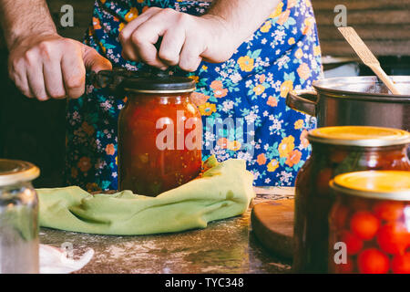 a man in a colored apron clogs tomatoes and Lecho sauce in glass jars in a farmhouse Stock Photo